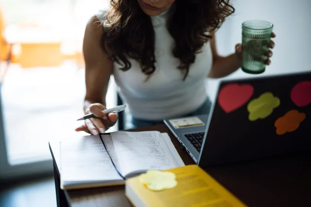 A woman sits at a desk working on a laptop while holding a green drink. She writes notes in a notebook. Sticky notes are placed on the laptop. The room is bright with sunlight. Small business CRM customer interviews: five prompts